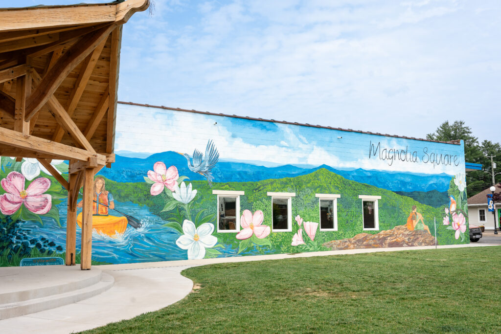 grassy outdoor space with stage and vibrant mural depicting outdoor recreation titled "Magnolia Square"